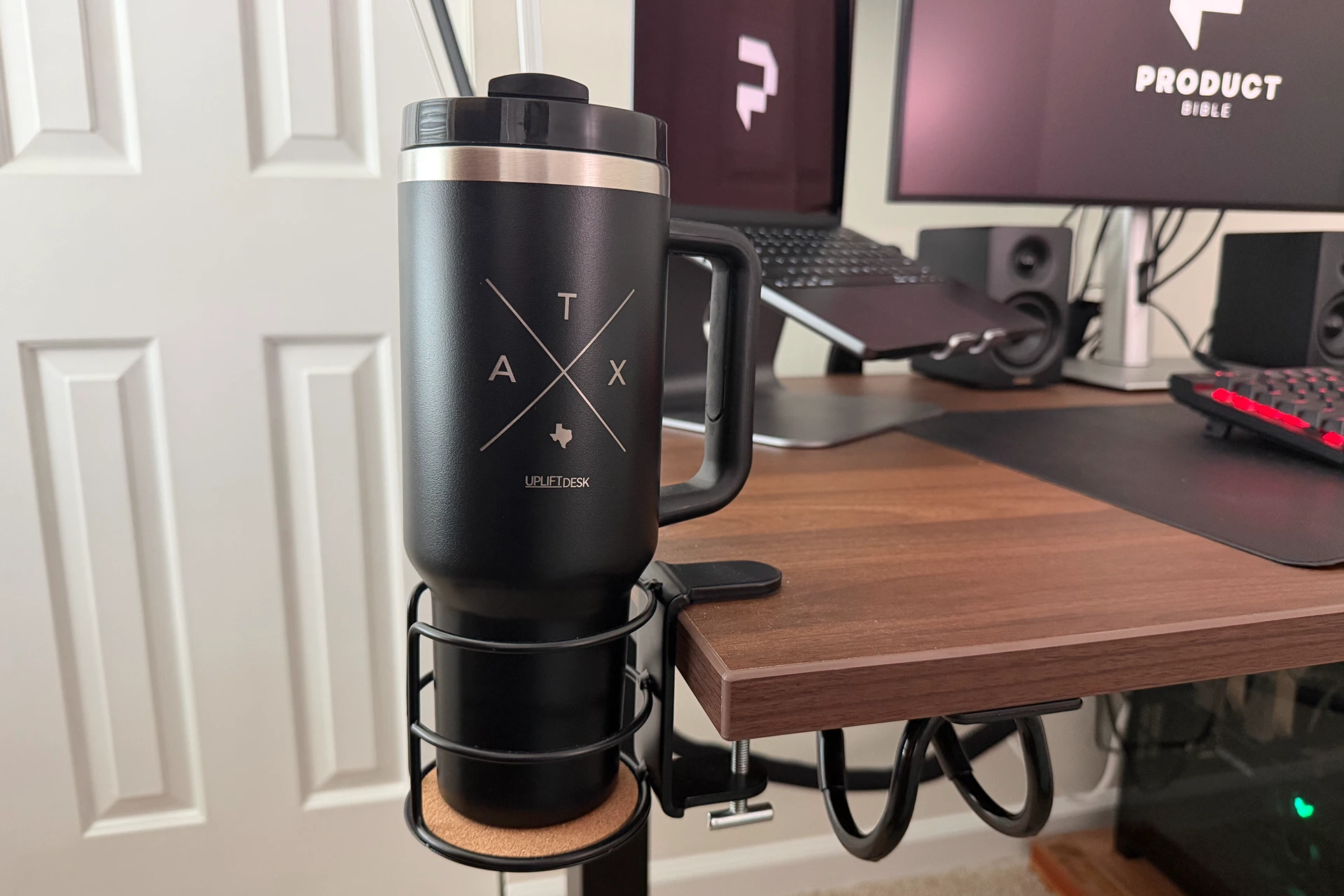 A close-up of the UPLIFT V3 standing desk cup holder mounted to the side of the walnut laminate desktop